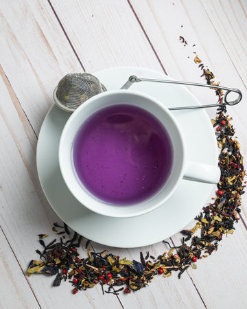 mug of purple tea with tea strainer and surrounded by the beautiful tea leaves used to brew